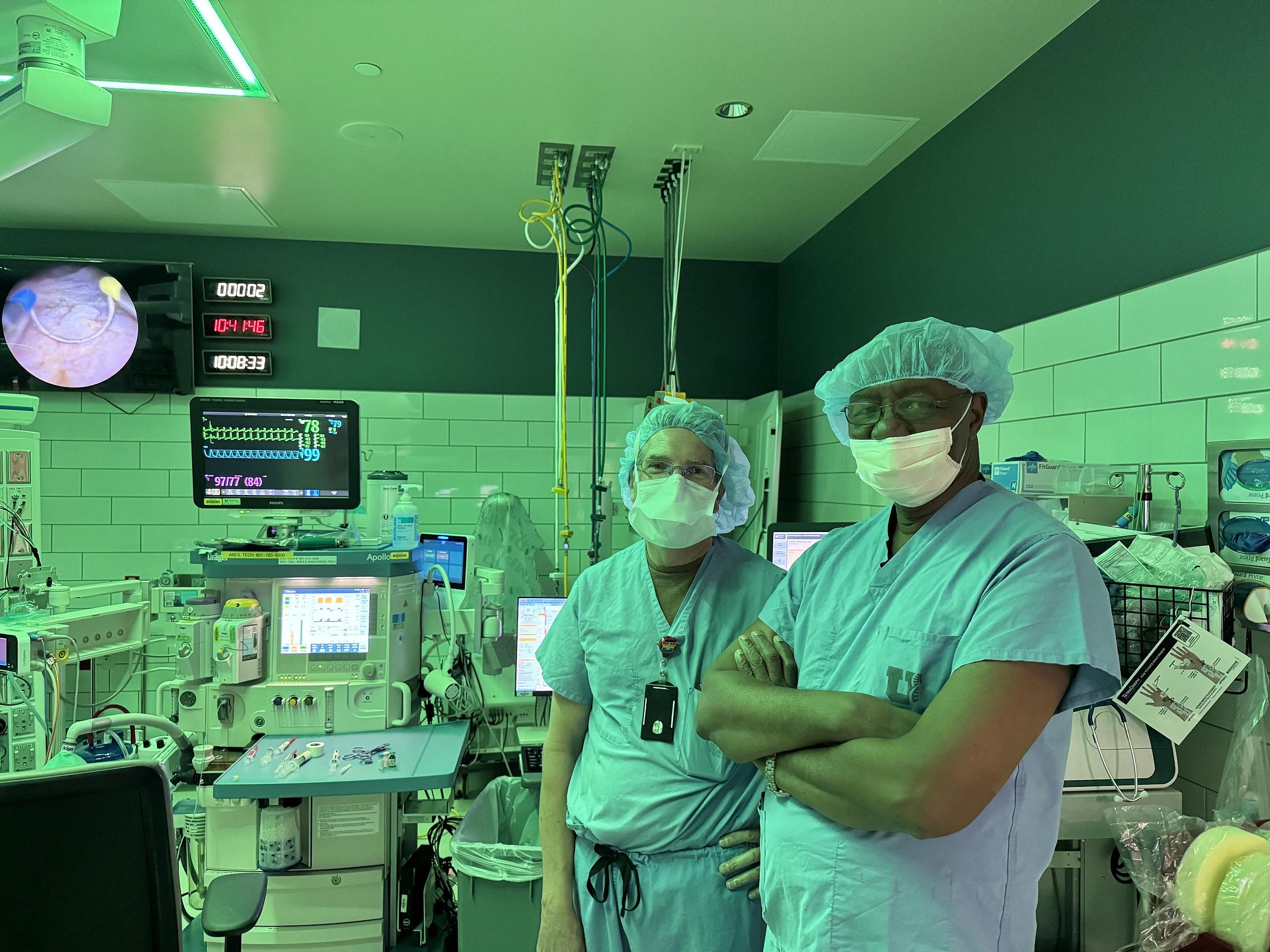Professor Baro and another doctor in an OR. The room is lit green with screens and medical equipment surrounds them.