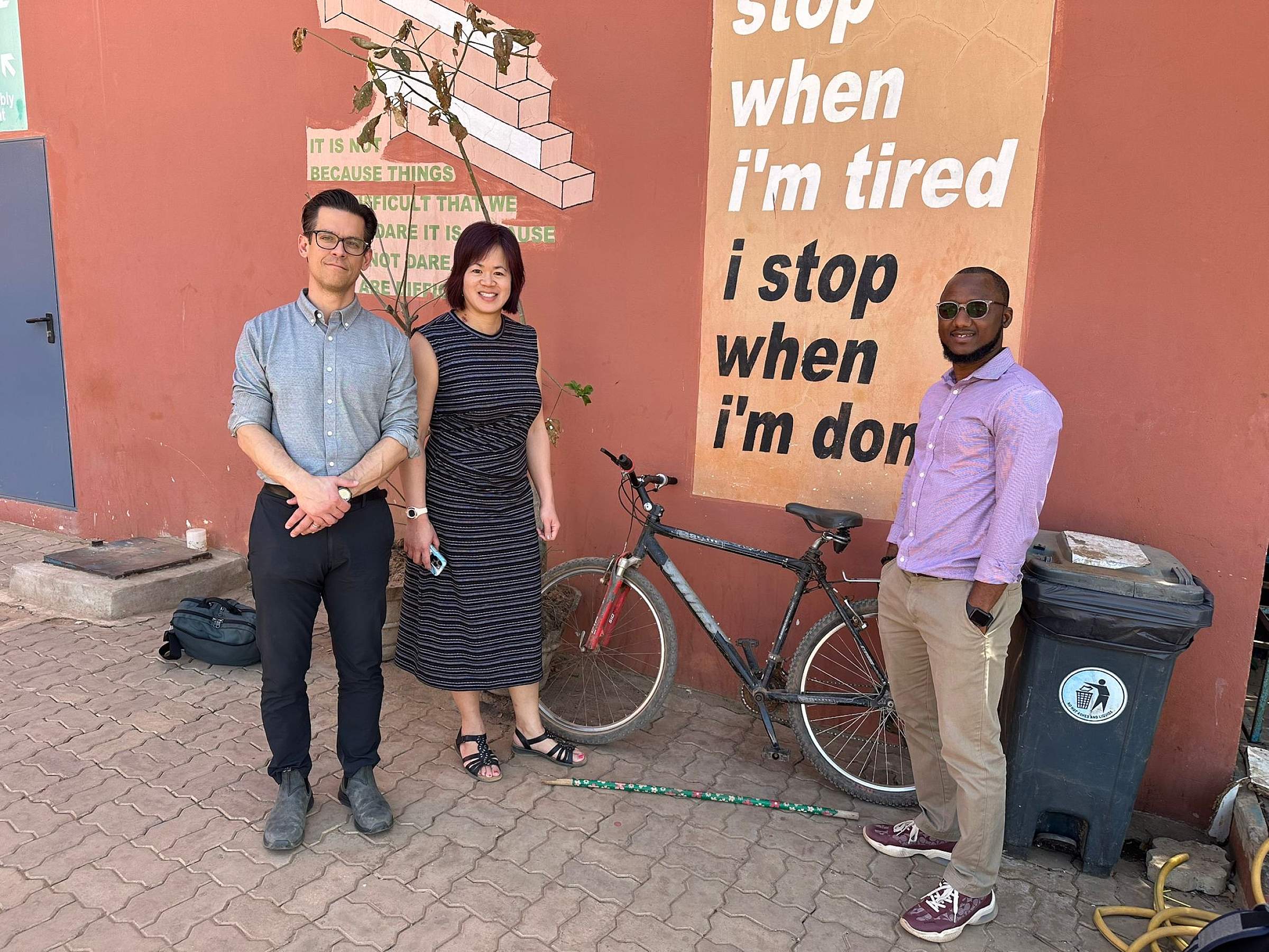 Drs. Scott Junkins, Candace Chang, and Kebbah Kah at Bundung Hospital. They smile at the camera in front of a mural that reads "I don't stop when I'm tired, I stop when I'm done."