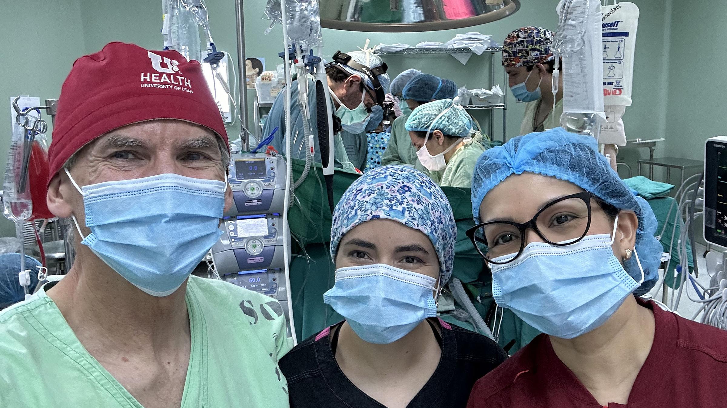 A group of 3 people in scrubs, caps, and masks taking a photo together in an OR. 