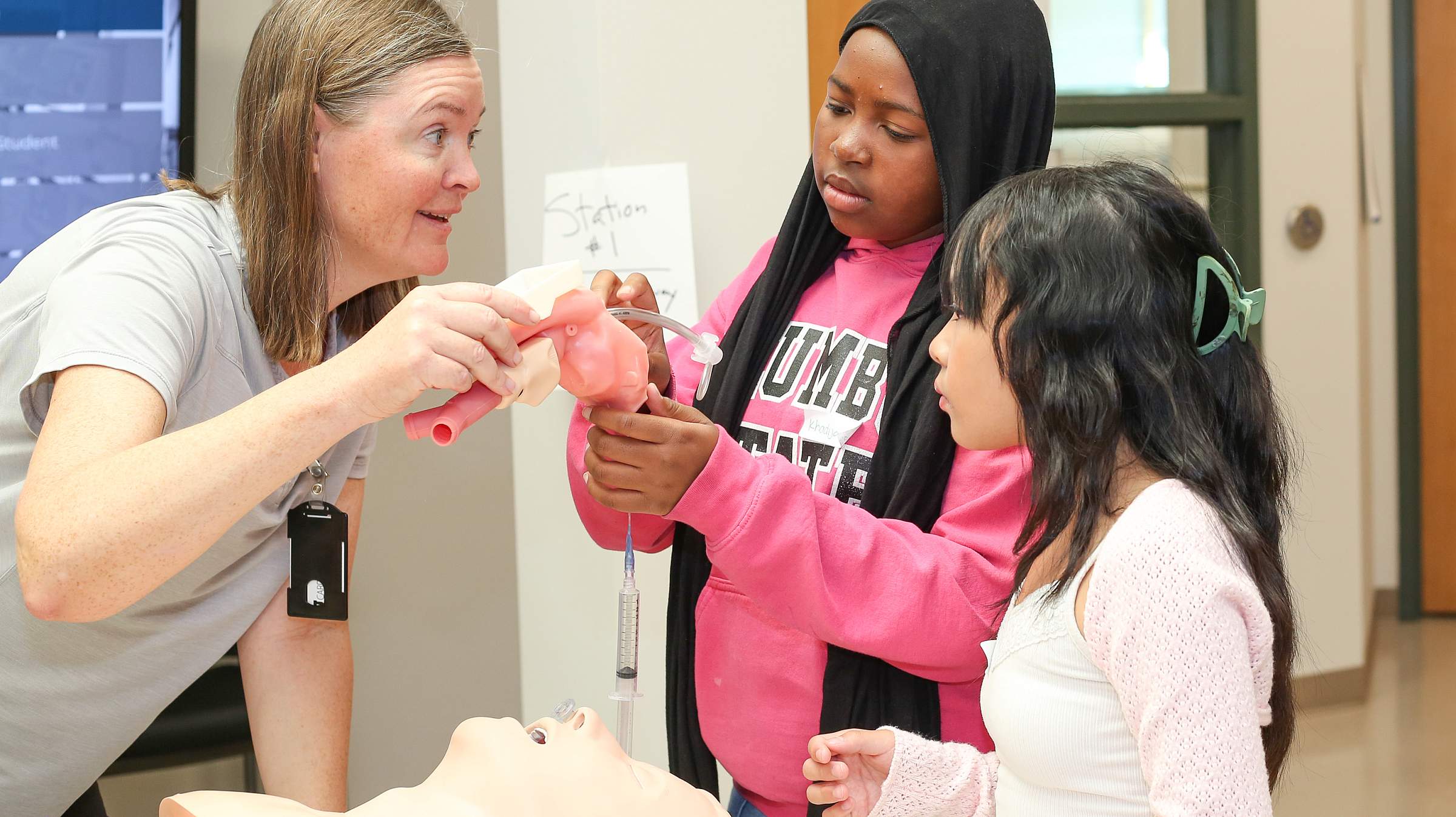 Two children and a doctor during an Imagine U school visit. The two children are on the right and the doctor is on the left. The gild in the center wears a pink hoodie and a black hair covering. The child on the far right wears a light pink sweater over a white shirt and has long black hair tied back with a green jaw clip. The doctor wears a grey shirt and holds a model of an human throat.